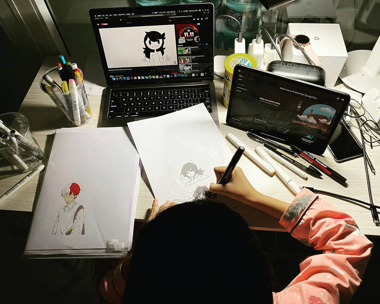 gallery-3 Artist sketching anime characters while watching a tutorial on a laptop, surrounded by artistic tools.