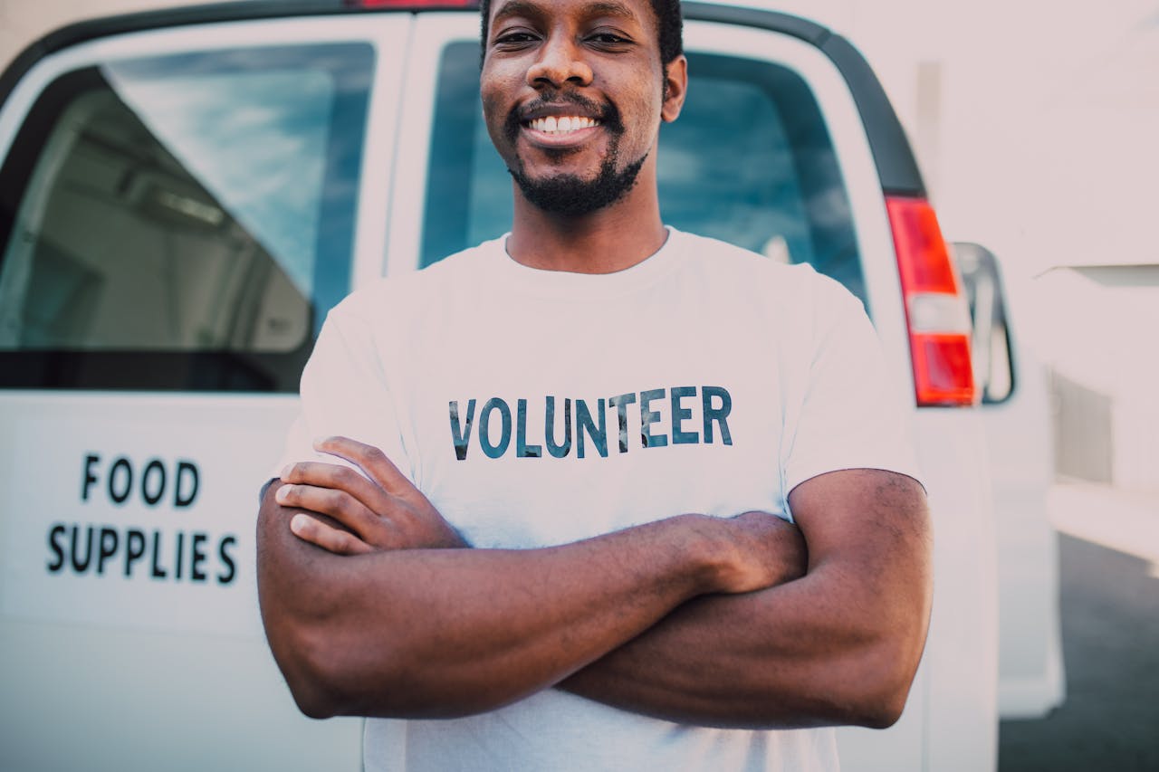 services-03 African American volunteer smiling in front of food supplies van with arms crossed, promoting community service.